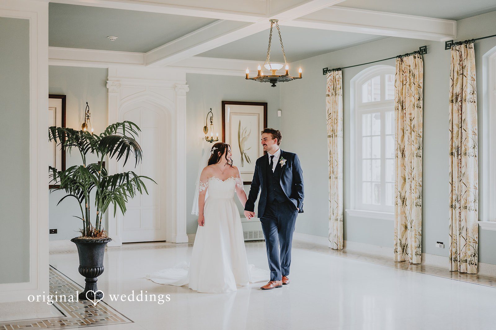 Bride and groom holding hands and standing in a bright hallway.
