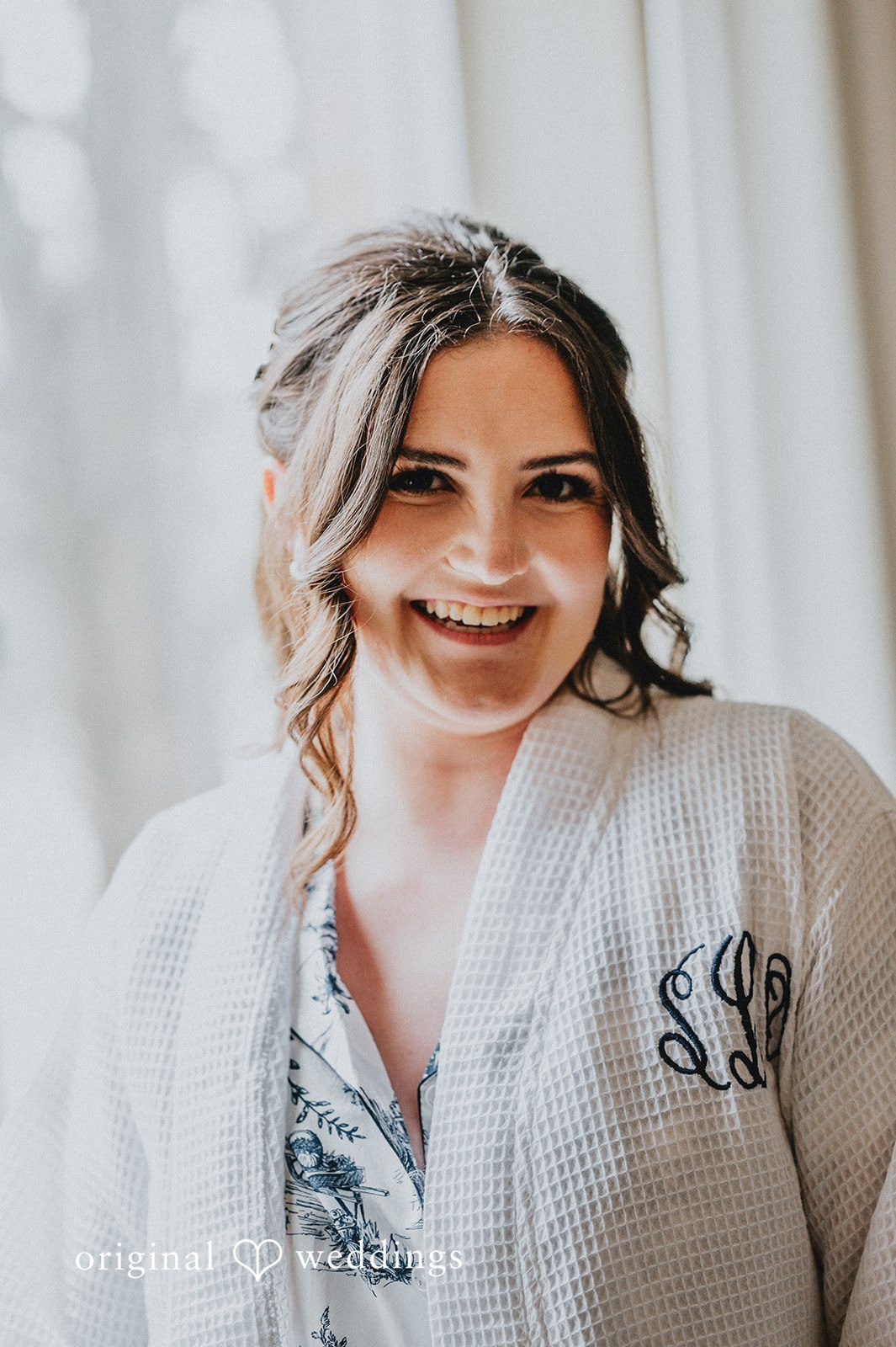 Portrait of a smiling bride wearing a white monogrammed robe while getting ready.