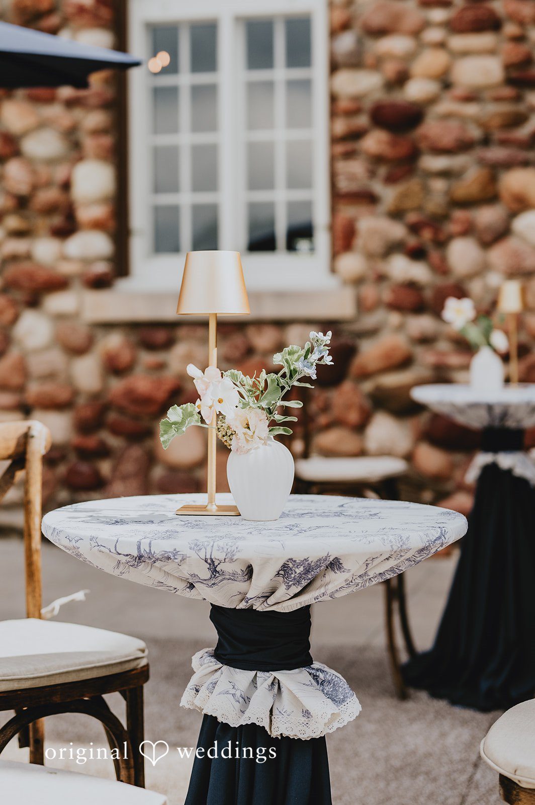 High-top cocktail table draped in white fabric next to a rustic stone wall.