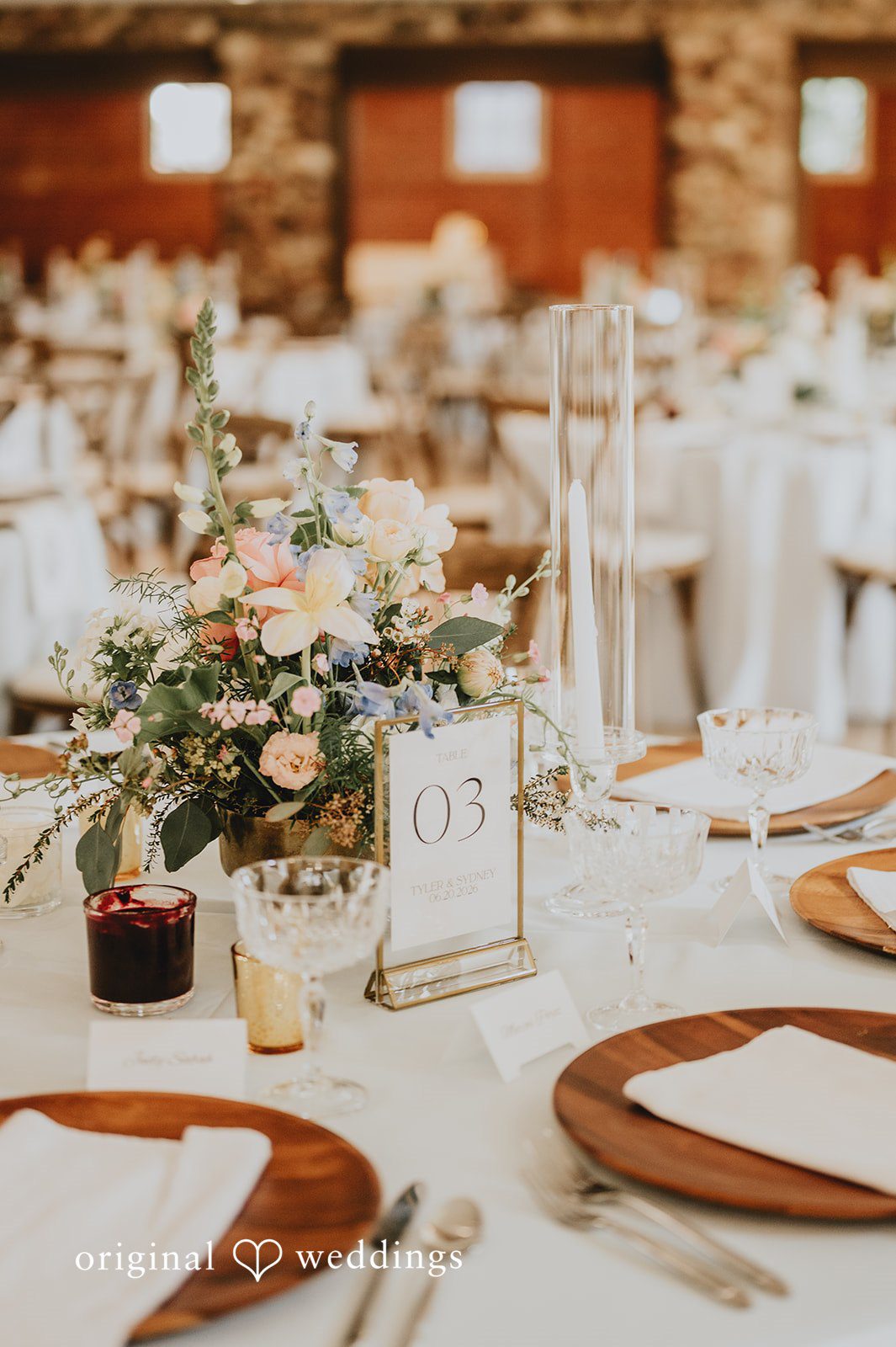 Close-up of a reception table setting featuring wooden chargers, white plates, and floral centerpieces.