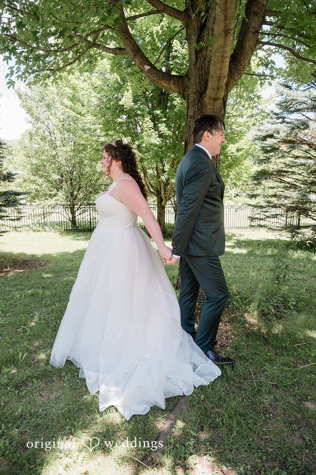 Carly + Seamus Bride and groom walking hand-in-hand near a line of trees.