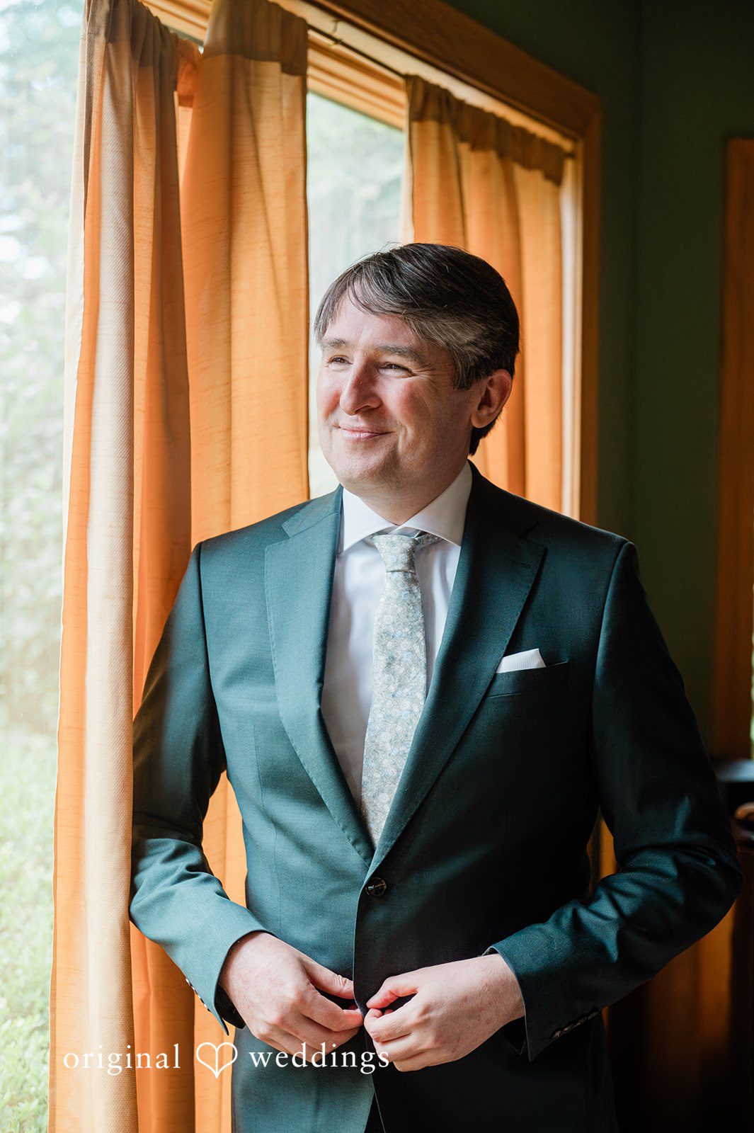Carly + Seamus Groom in a green suit smiling while adjusting his jacket.