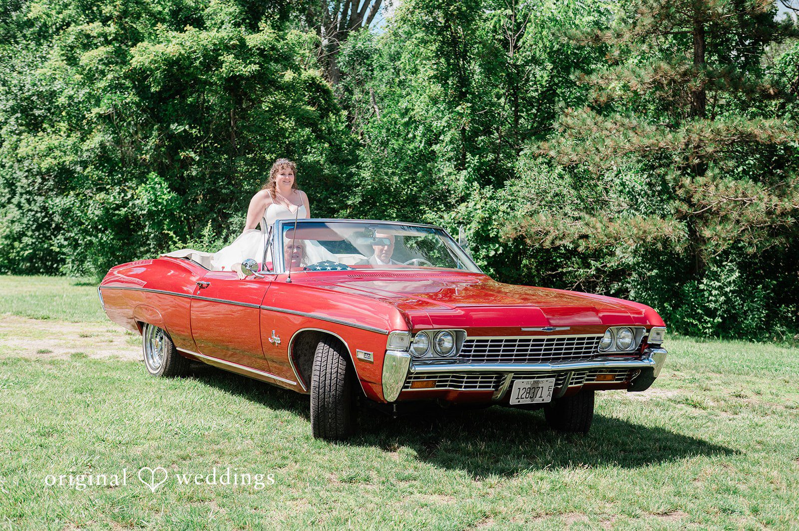 Carly + Seamus Bride standing in the back seat of a moving red convertible car on a lawn.