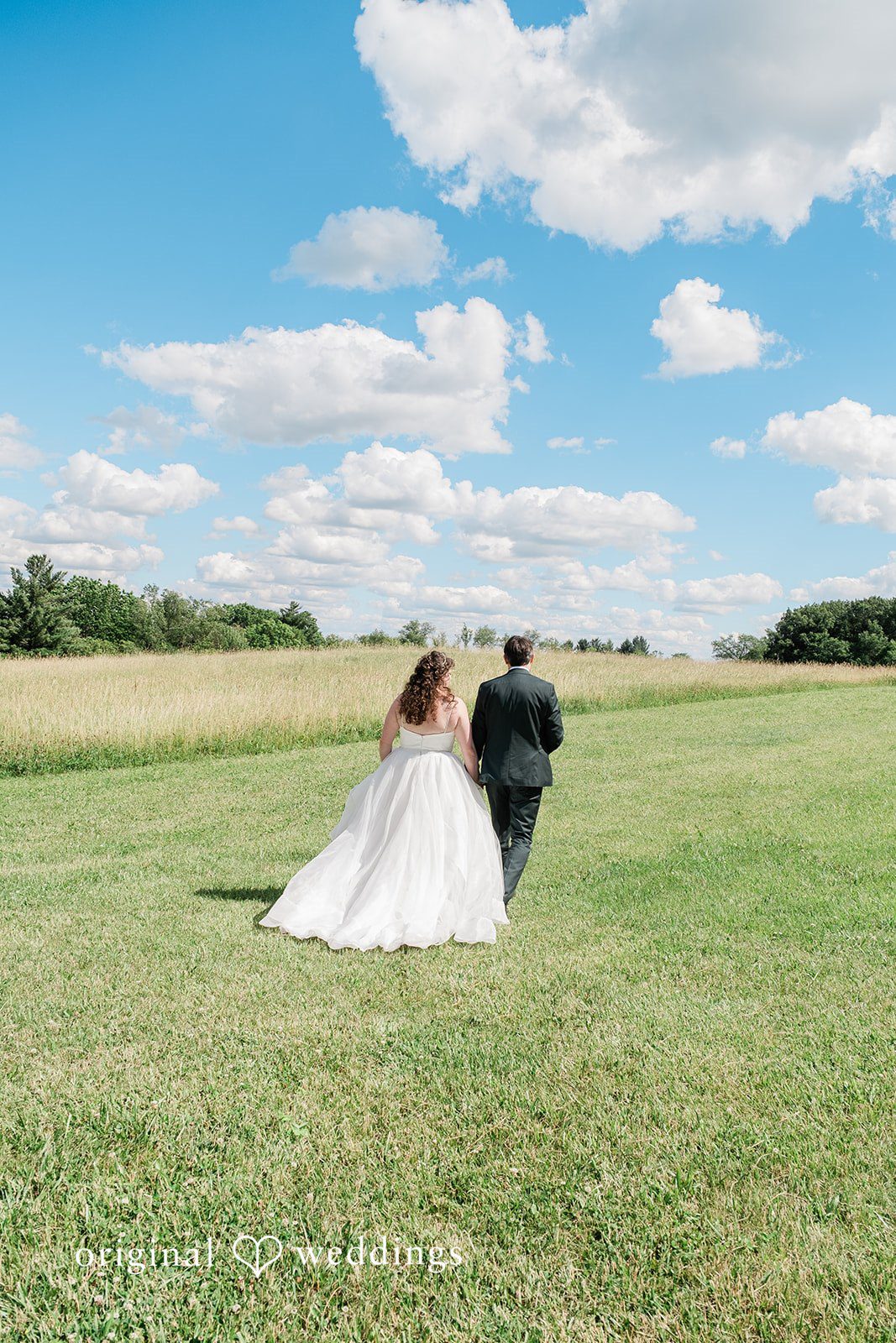 Carly + Seamus Wide shot of a bride and groom walking across a large grassy field.