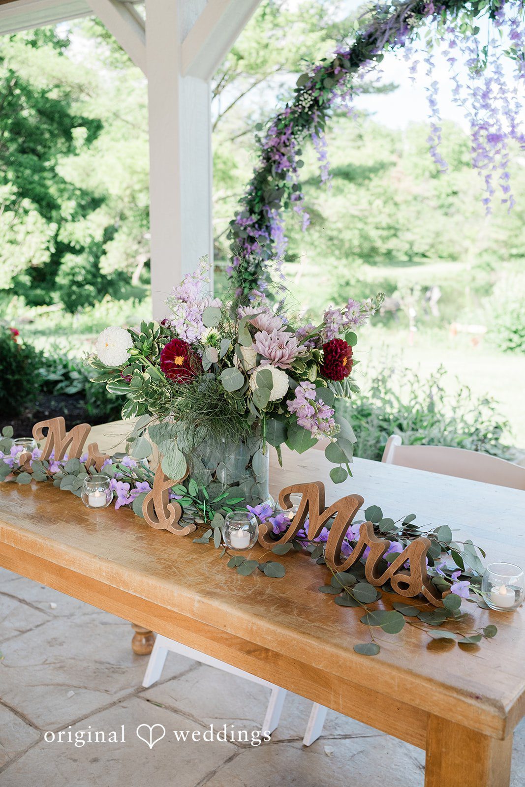 Carly + Seamus Close-up of a rustic wooden banquet table decorated with greenery and floral arrangements.