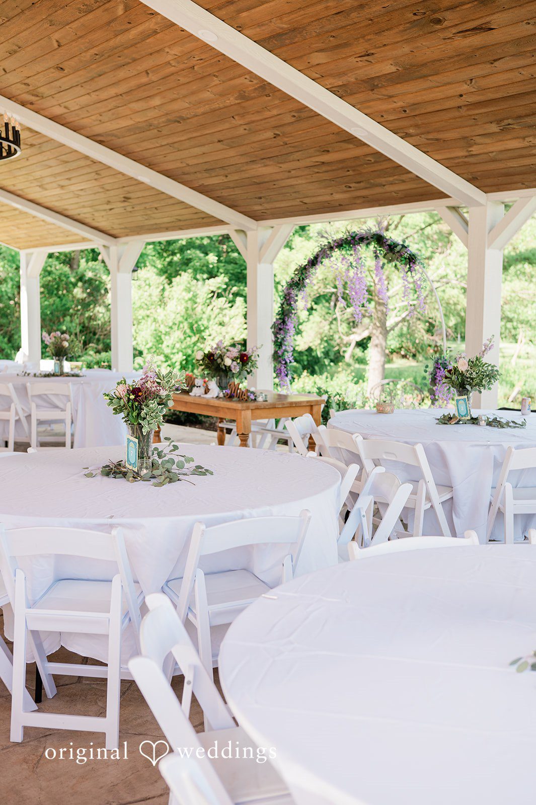 Carly + Seamus Round dining tables set with white tablecloths and centerpieces under a wooden pavilion.
