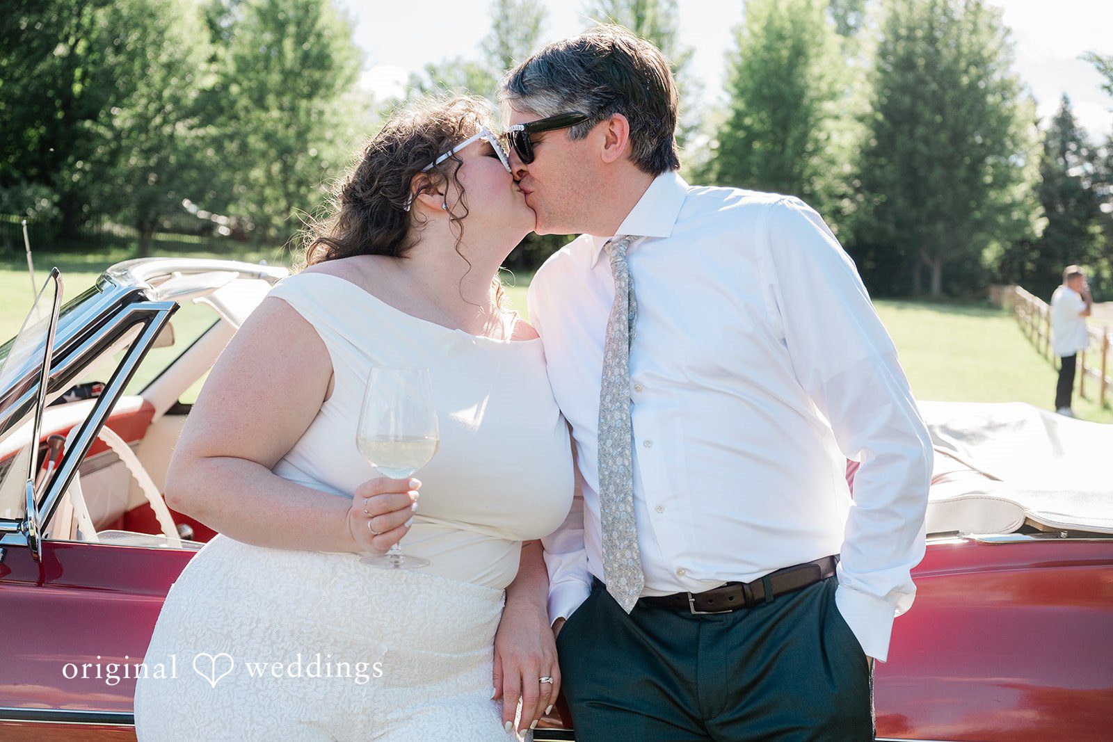Carly + Seamus Bride in a white jumpsuit and groom kissing in front of a red convertible.