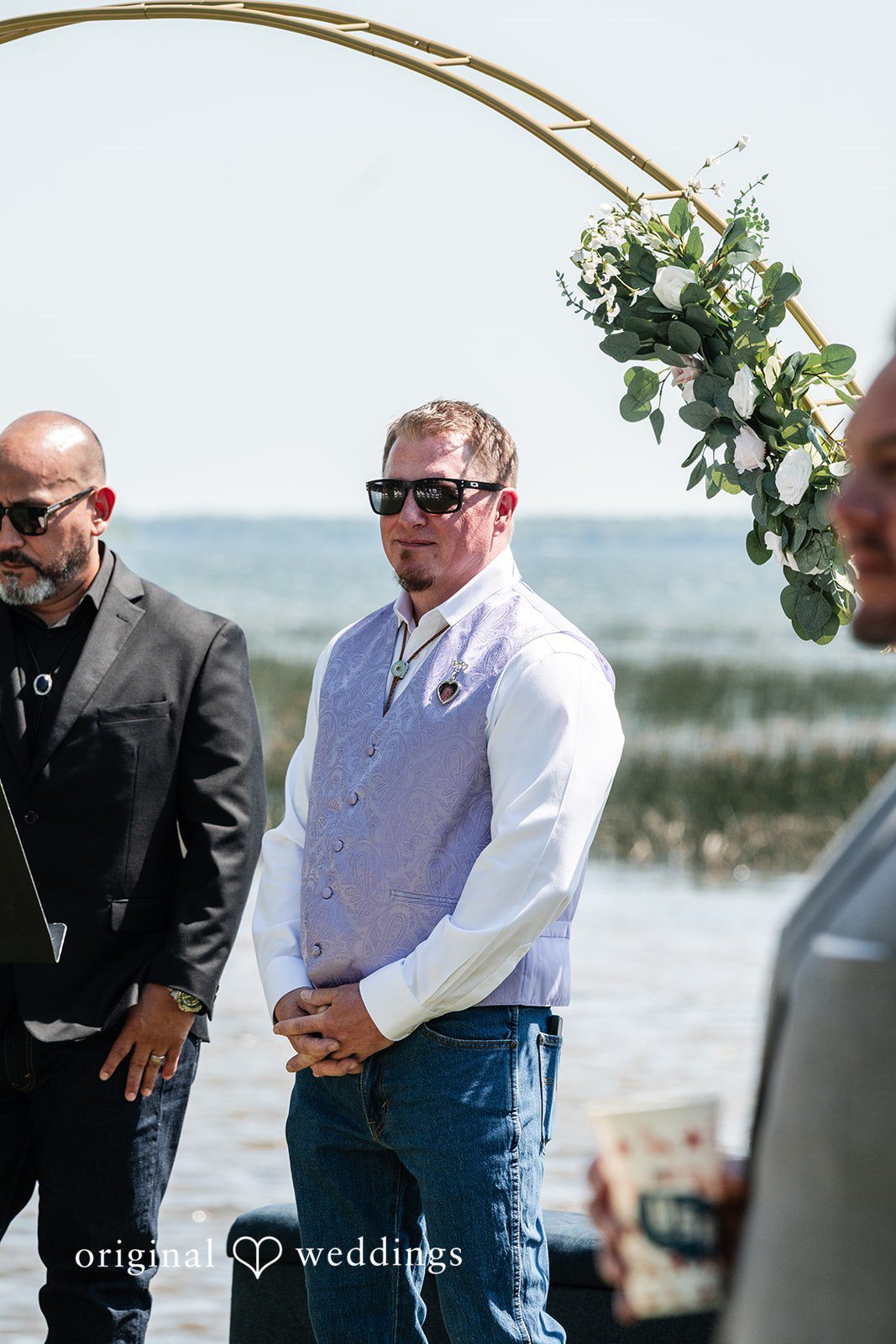 A groom wearing a white shirt and vest standing near a waterfront ceremony.