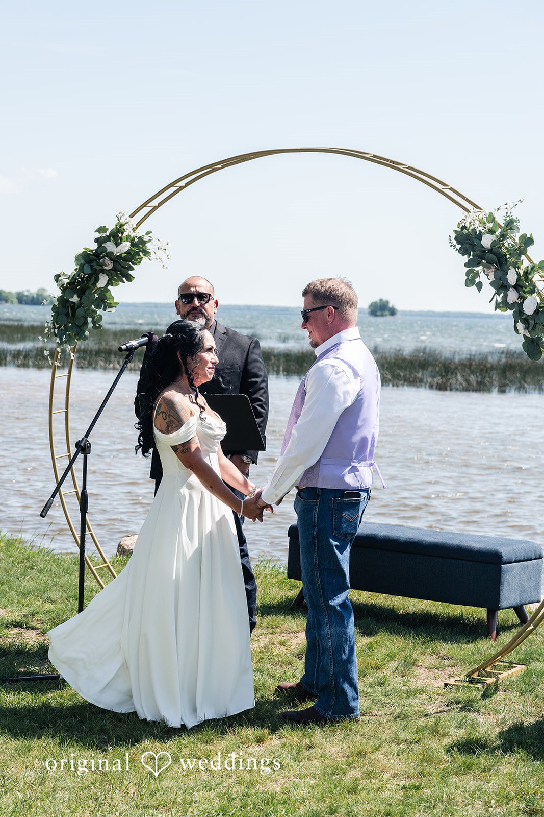 A bride and groom standing under a circular arch during a waterfront ceremony.