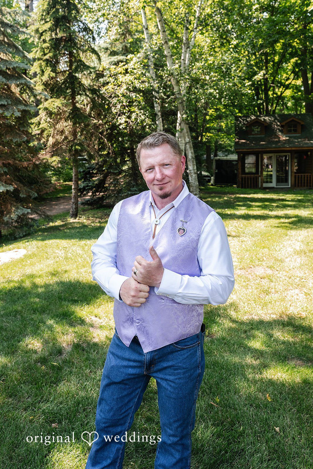 A man wearing a light purple vest and jeans smiling on a grassy field.