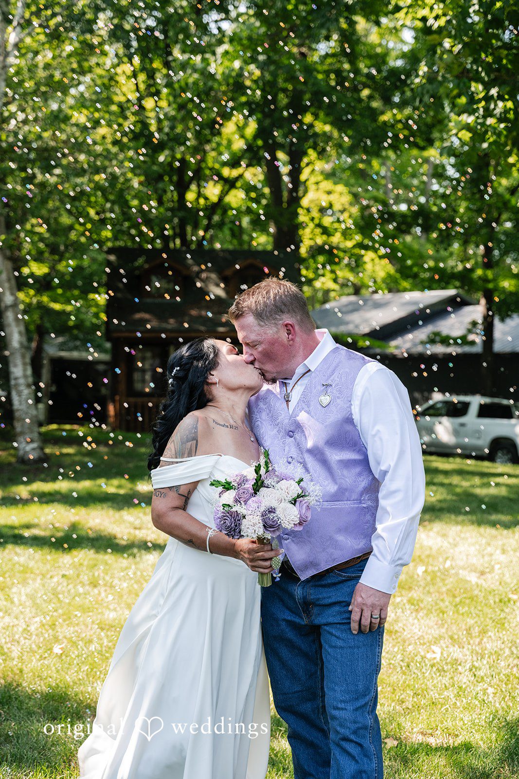 A groom and bride kissing while standing outdoors surrounded by trees.