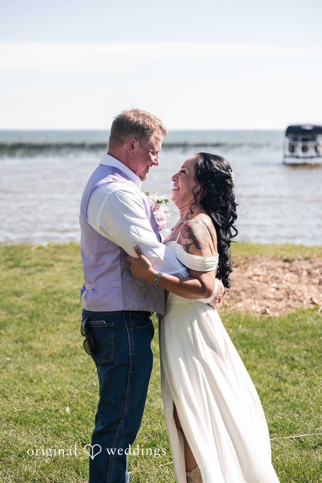 A groom hugging a bride from behind with a lake and clear sky in the background.