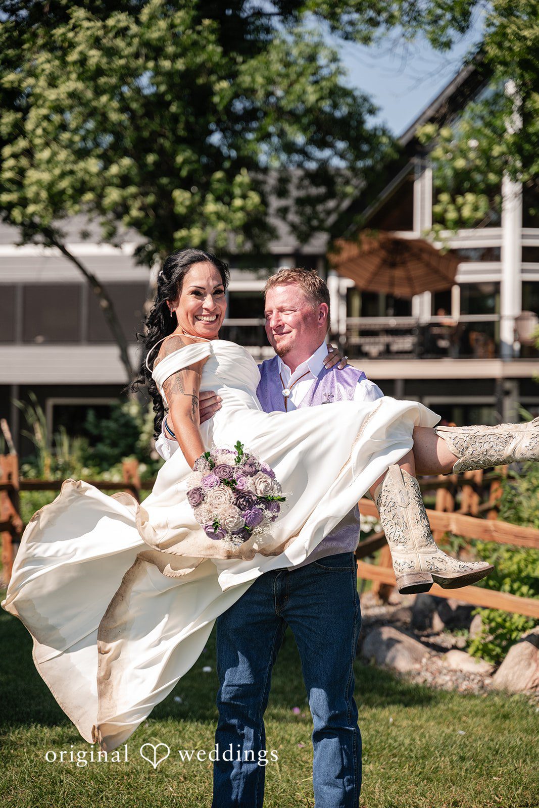 A groom lifting a bride in a white dress up in his arms outside a rustic building.
