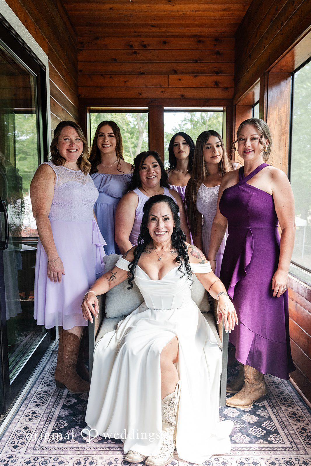 A bride seated in a chair surrounded by bridesmaids in matching purple dresses.