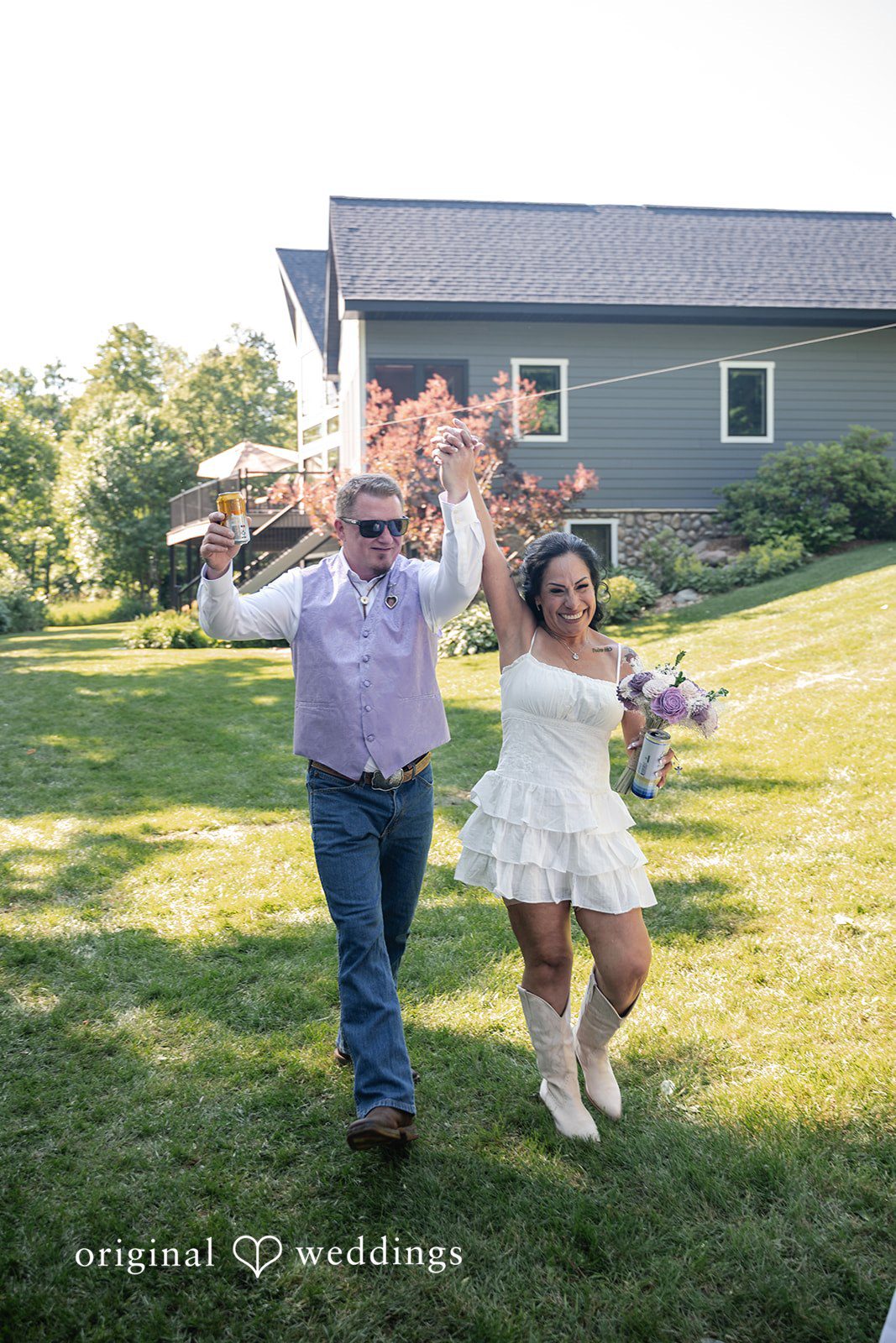 A groom and a bride cheering with raised arms while walking down a grassy slope.