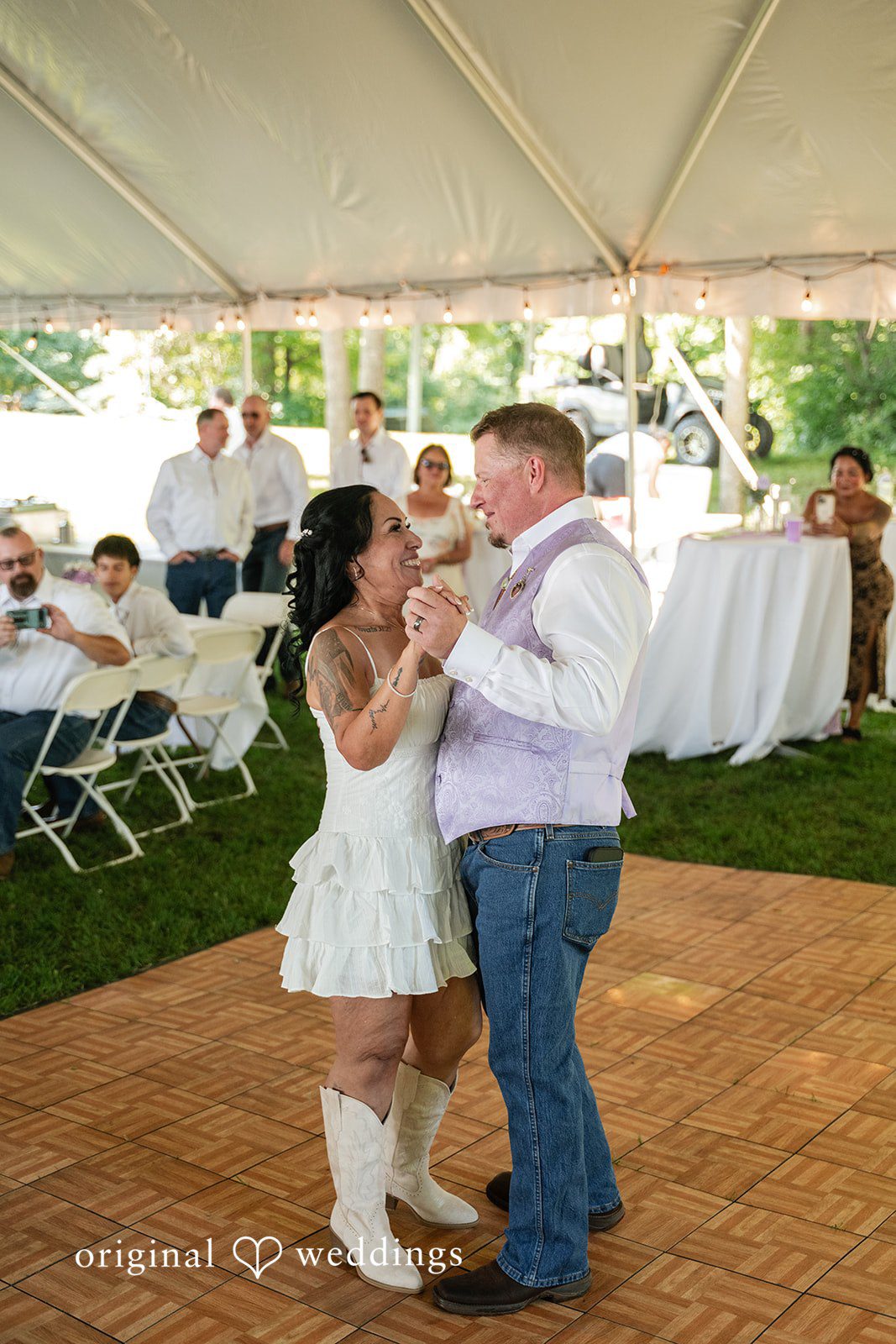 A smiling bride and a groom in a vest dance together as a happy couple on a wooden dance floor.