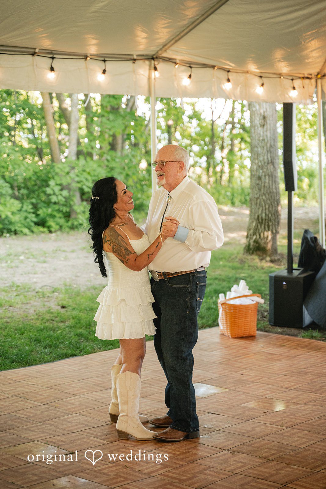 A smiling bride in a short white dress and cowboy boots dances with an older man in a white shirt and jeans on a wooden dance floor.