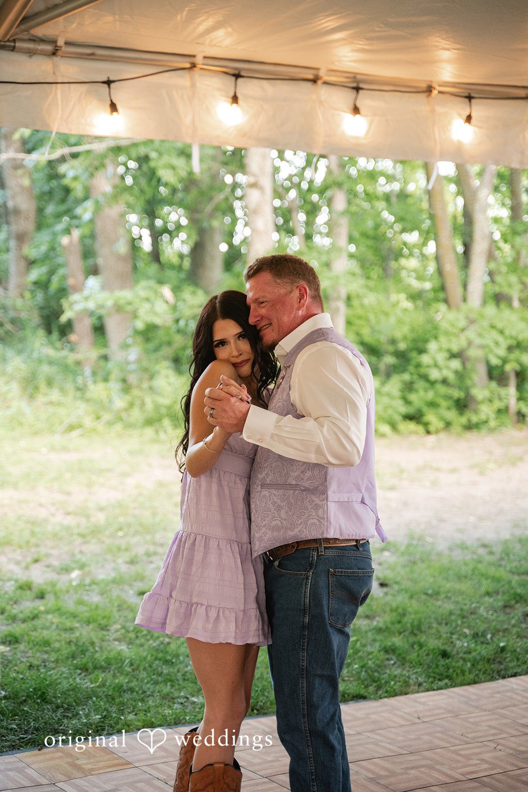 A groom and a woman dancing closely together on a wooden floor under string lights.