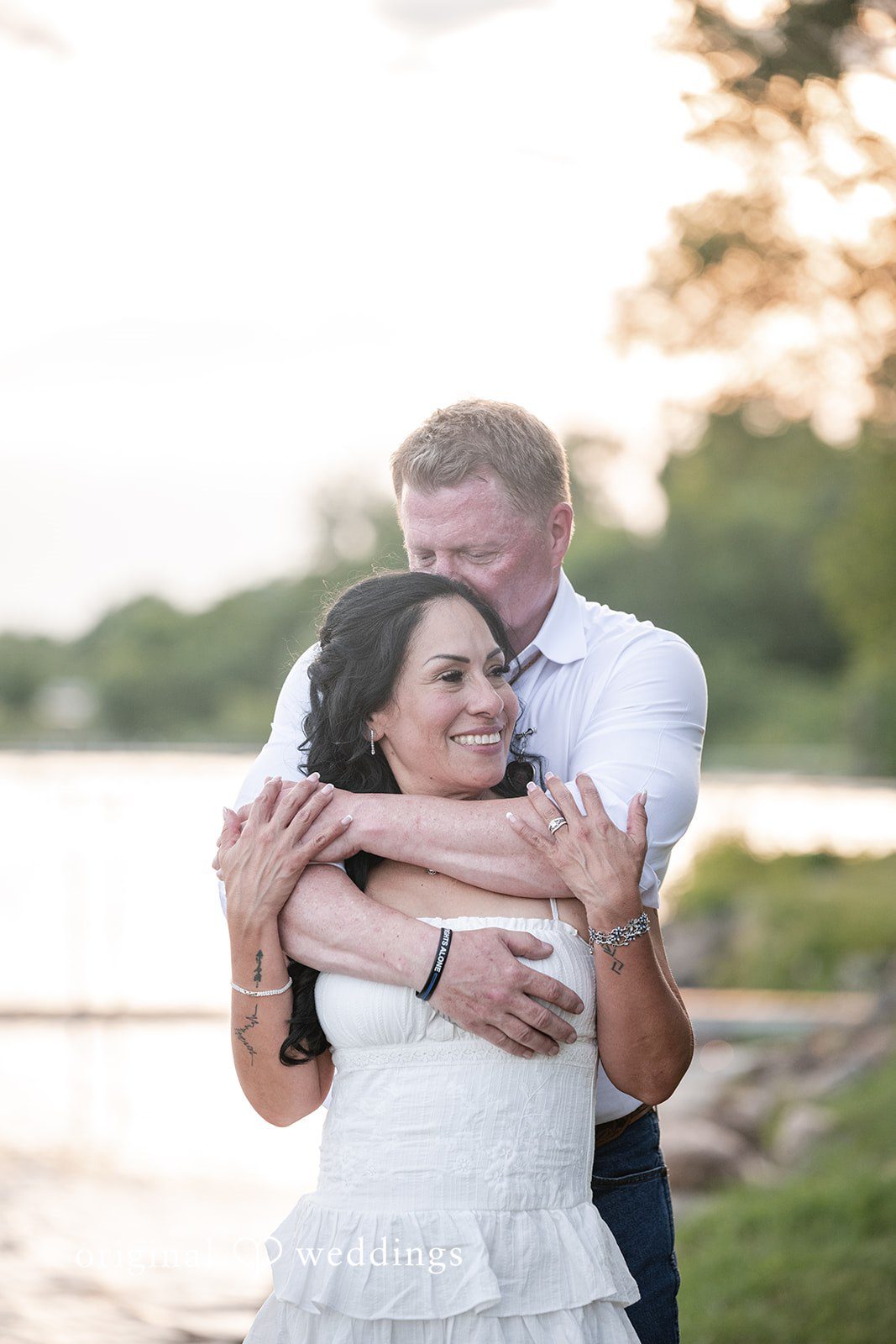 A groom hugging a smiling bride tightly from behind in a close-up portrait.