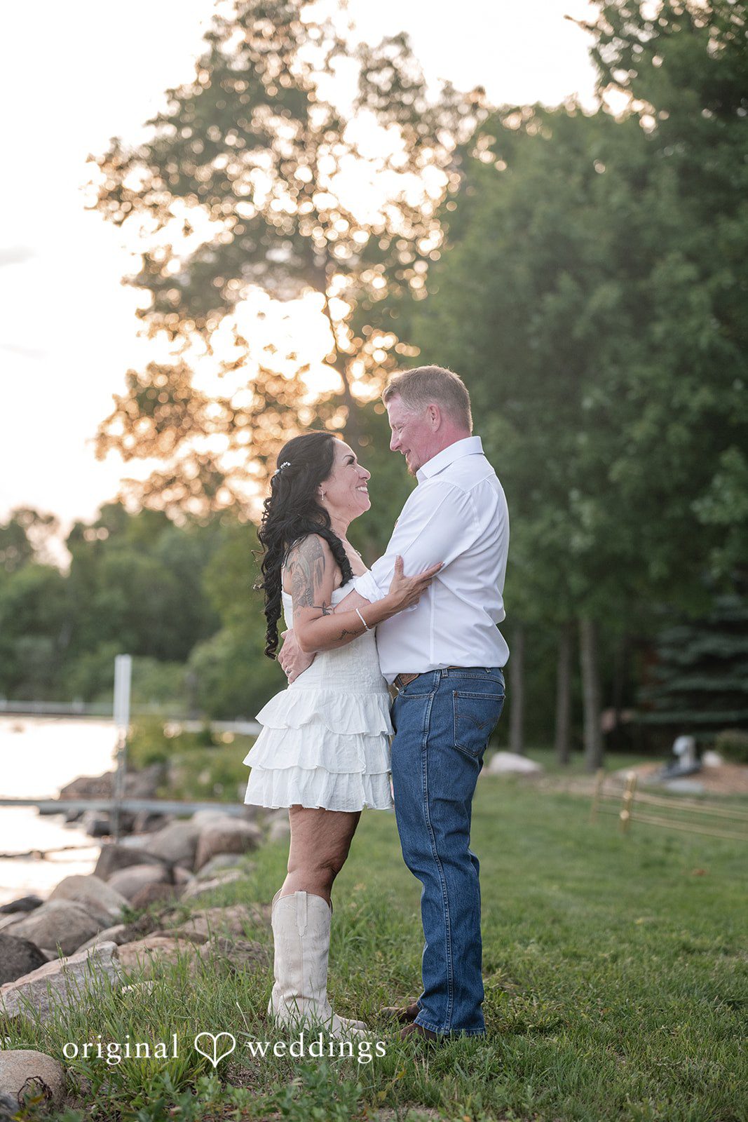 A groom and a bride standing face-to-face on a lakeside shore during sunset.