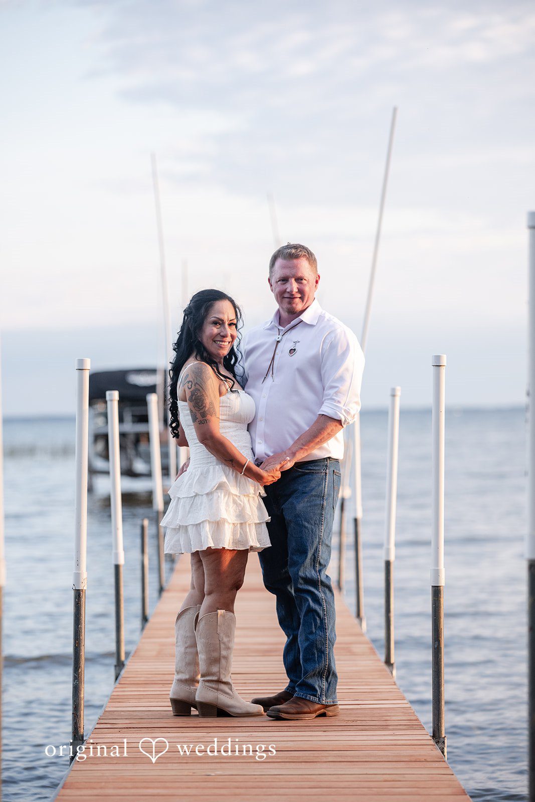 Couple holding hands while standing on a wooden dock extending into a lake.