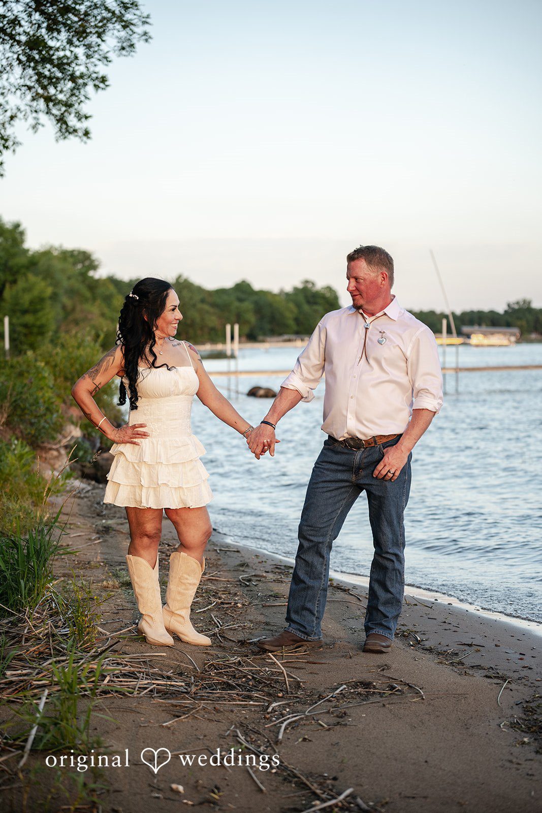 Couple holding hands on a rocky beach with water behind them.