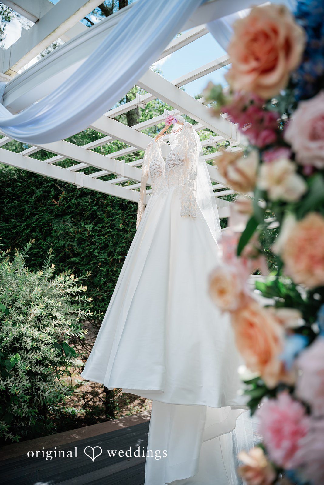 Jessica + Patrick A wedding dress hanging elegantly under a garden pergola adorned with pink flowers.