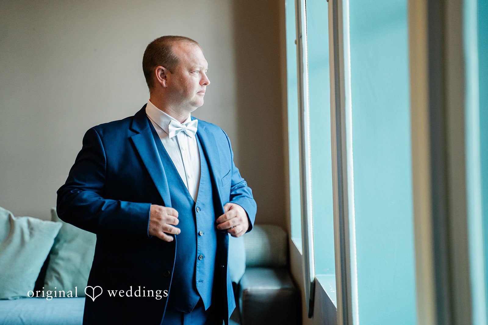 Jessica + Patrick A man in a blue suit adjusts his jacket while looking out a bright window.