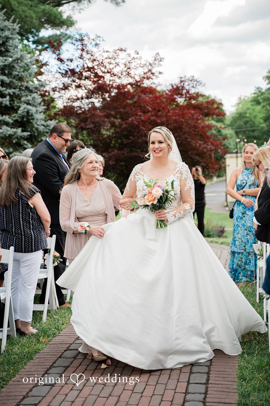 Jessica + Patrick A bride in a large ballgown walks down a brick aisle between rows of guests.