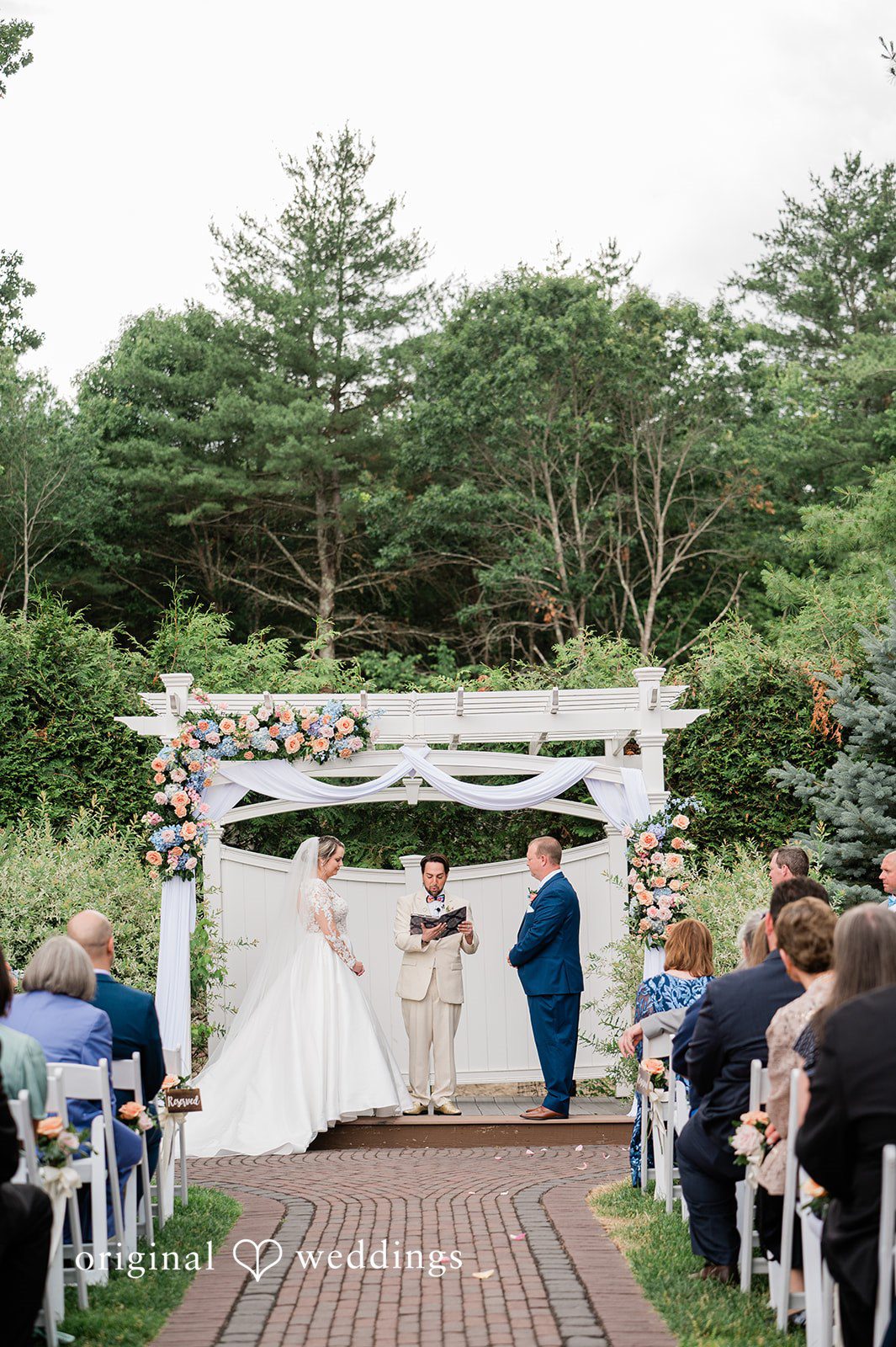 Jessica + Patrick An elevated view of a ceremony at a white wooden arbor with guests seated on both sides.