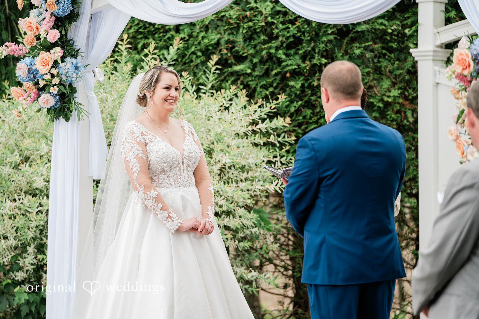 Jessica + Patrick A bride looks at her groom while standing at the altar framed by green foliage.