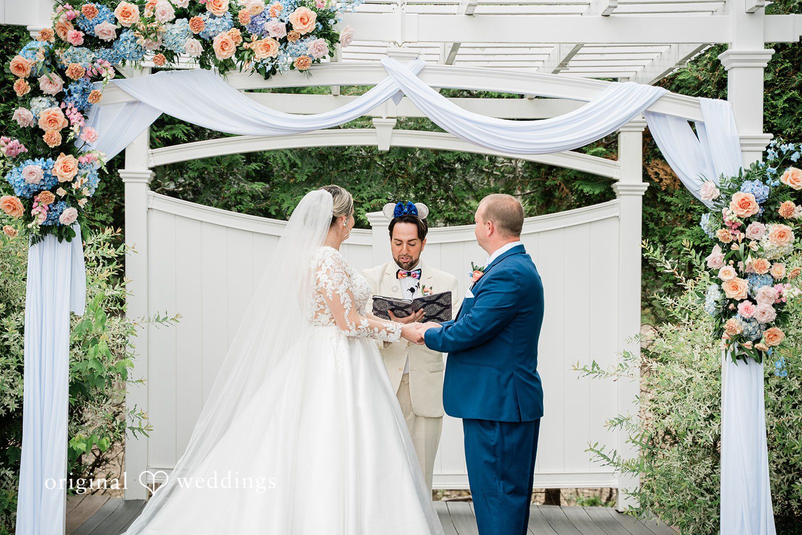 Jessica + Patrick A bride and groom hold hands and exchange vows in front of a white arbor.