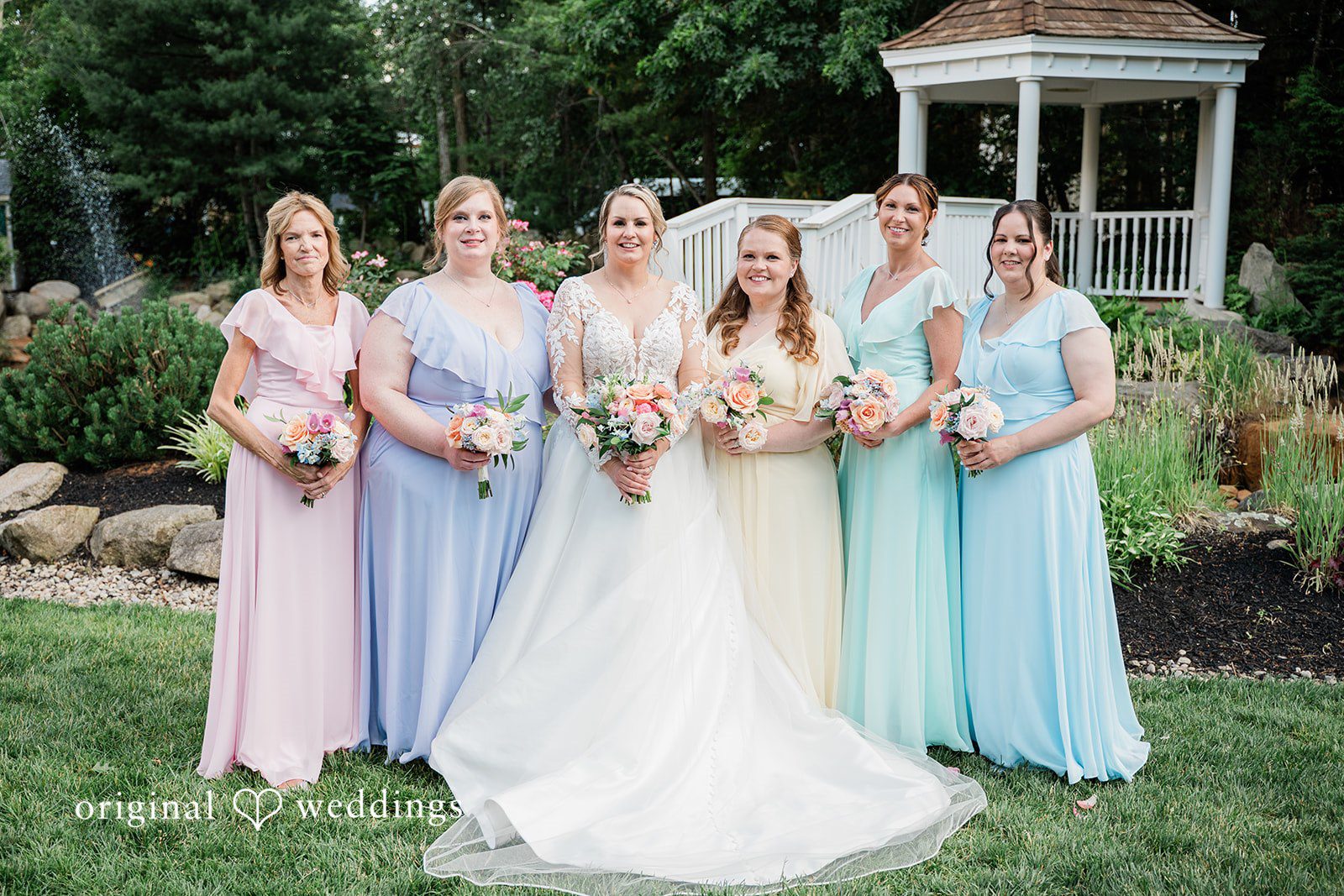 Jessica + Patrick Bride and Bridesmaid in pastel dresses stand together holding bouquets in a garden.