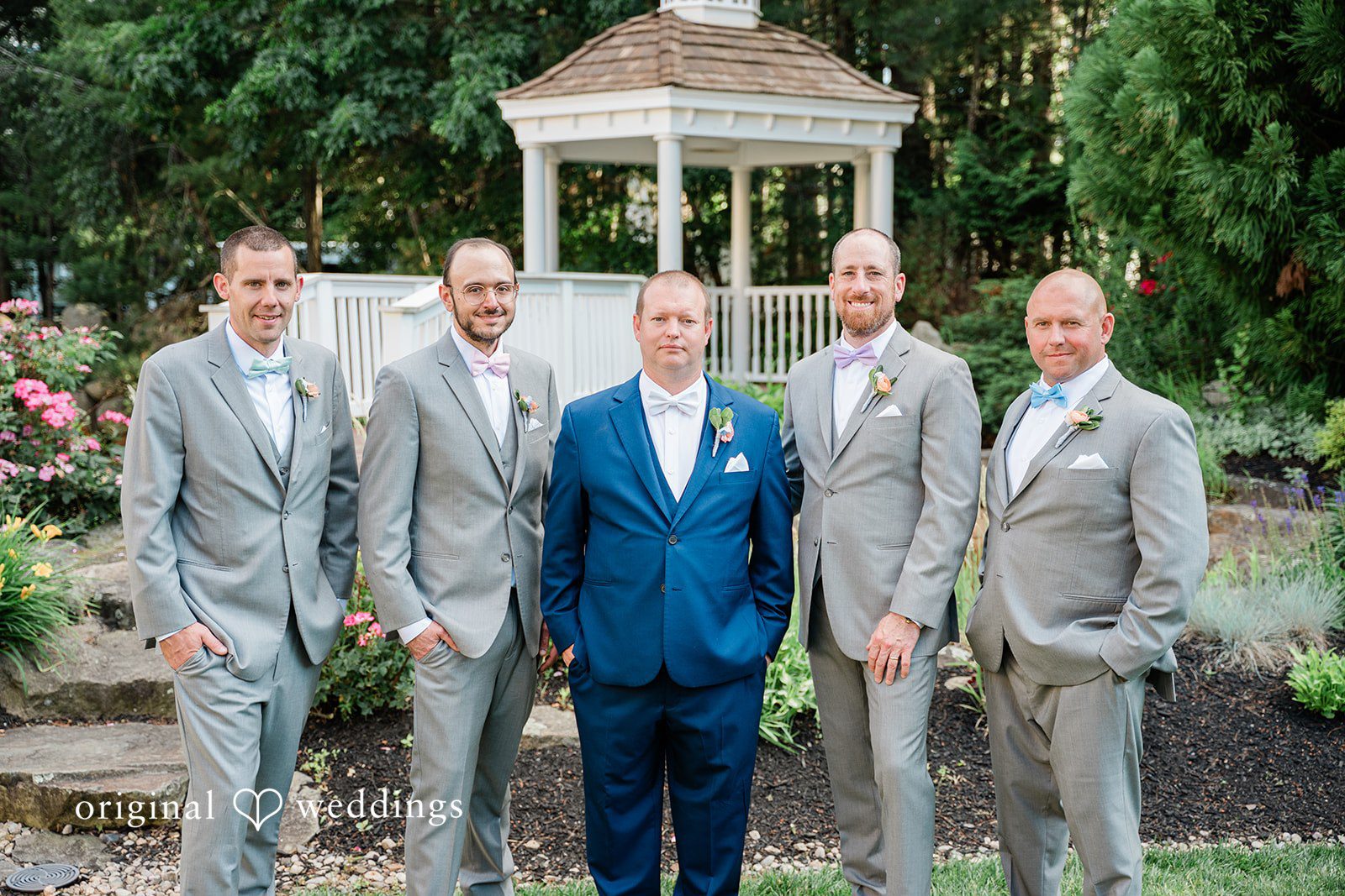Jessica + Patrick Grooms and groomsmen in matching suits stand in a row smiling in front of a white gazebo.
