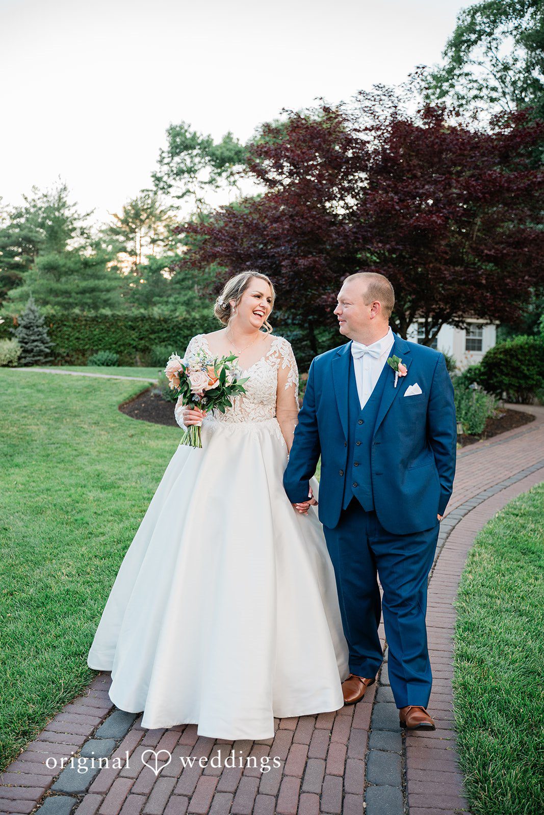 Jessica + Patrick A bride and groom walk hand-in-hand down a paved brick path.