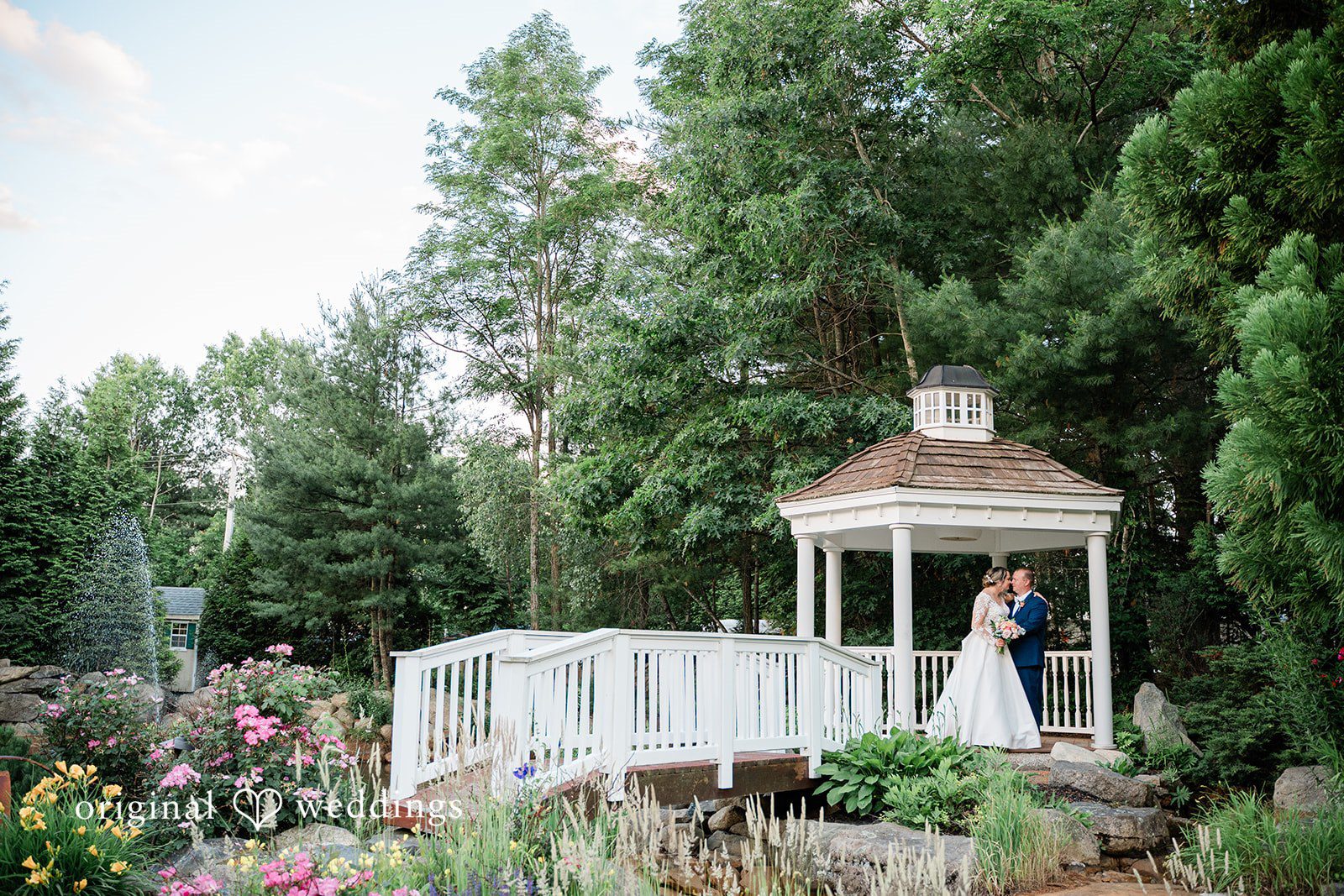 Jessica + Patrick A bride and groom stand together inside a white garden gazebo surrounded by trees.