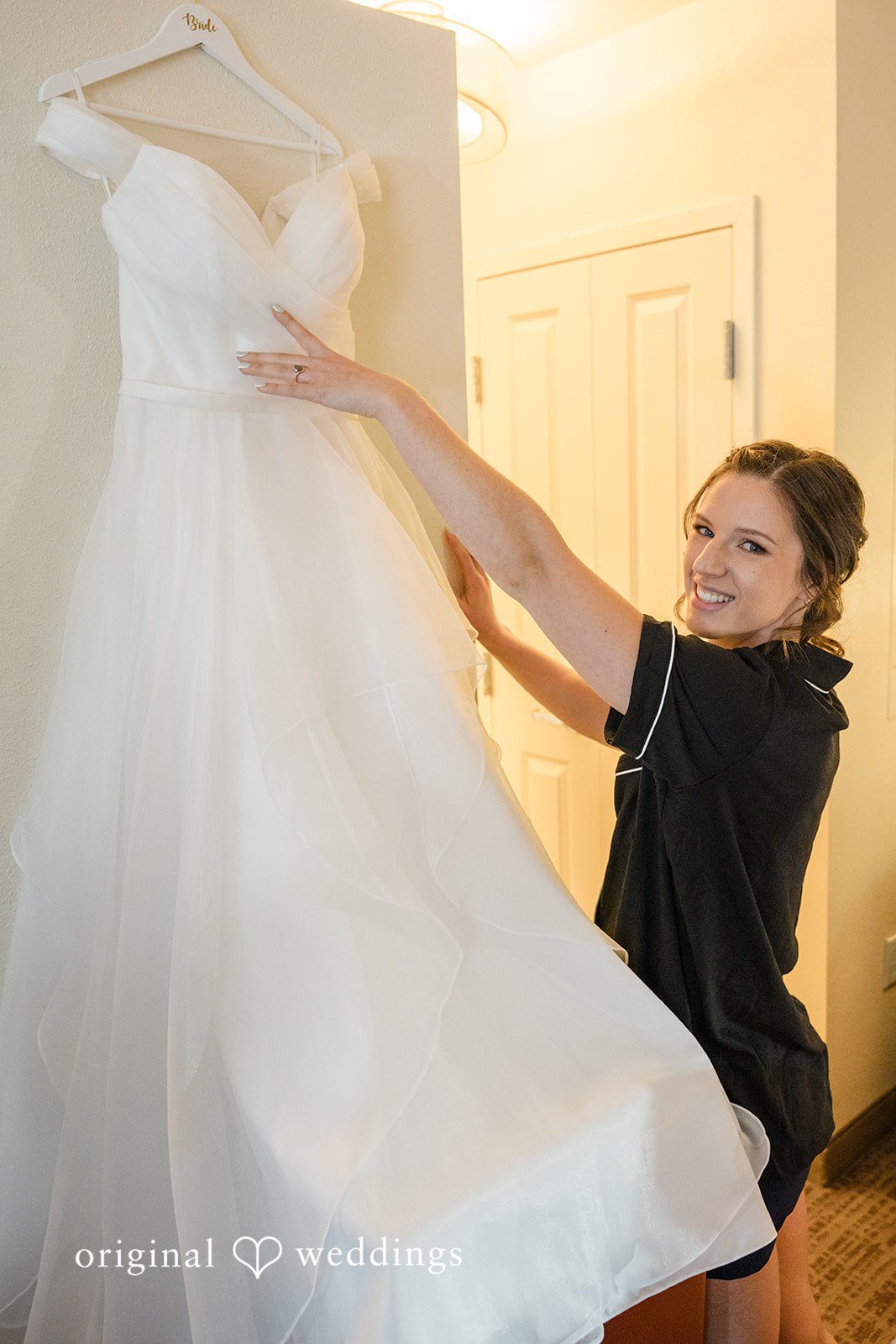Emily + Maxwell A woman adjusting a white wedding dress hanging on a wall.