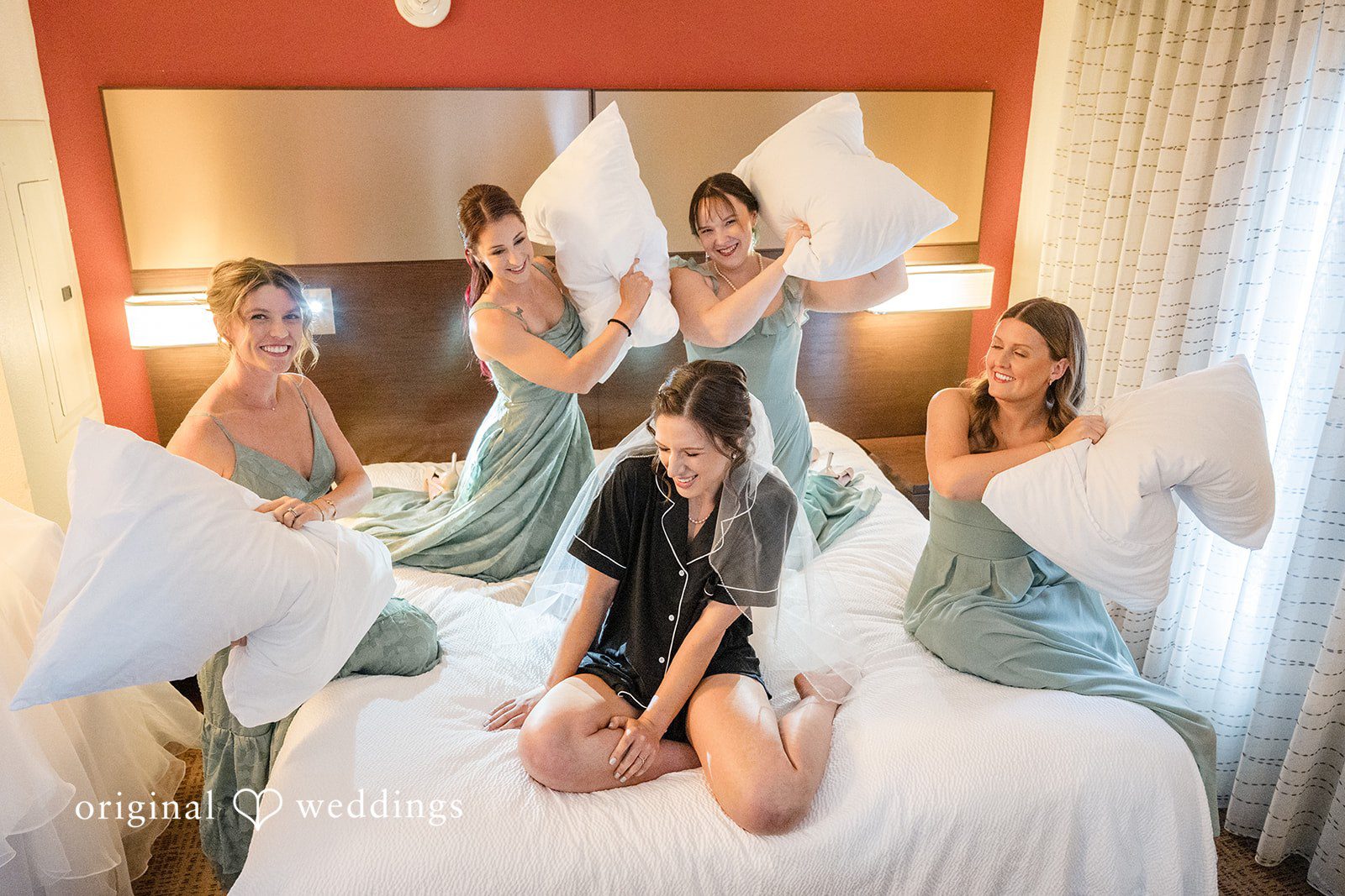 Emily + Maxwell A bride and her bridesmaids having a playful pillow fight on a bed.