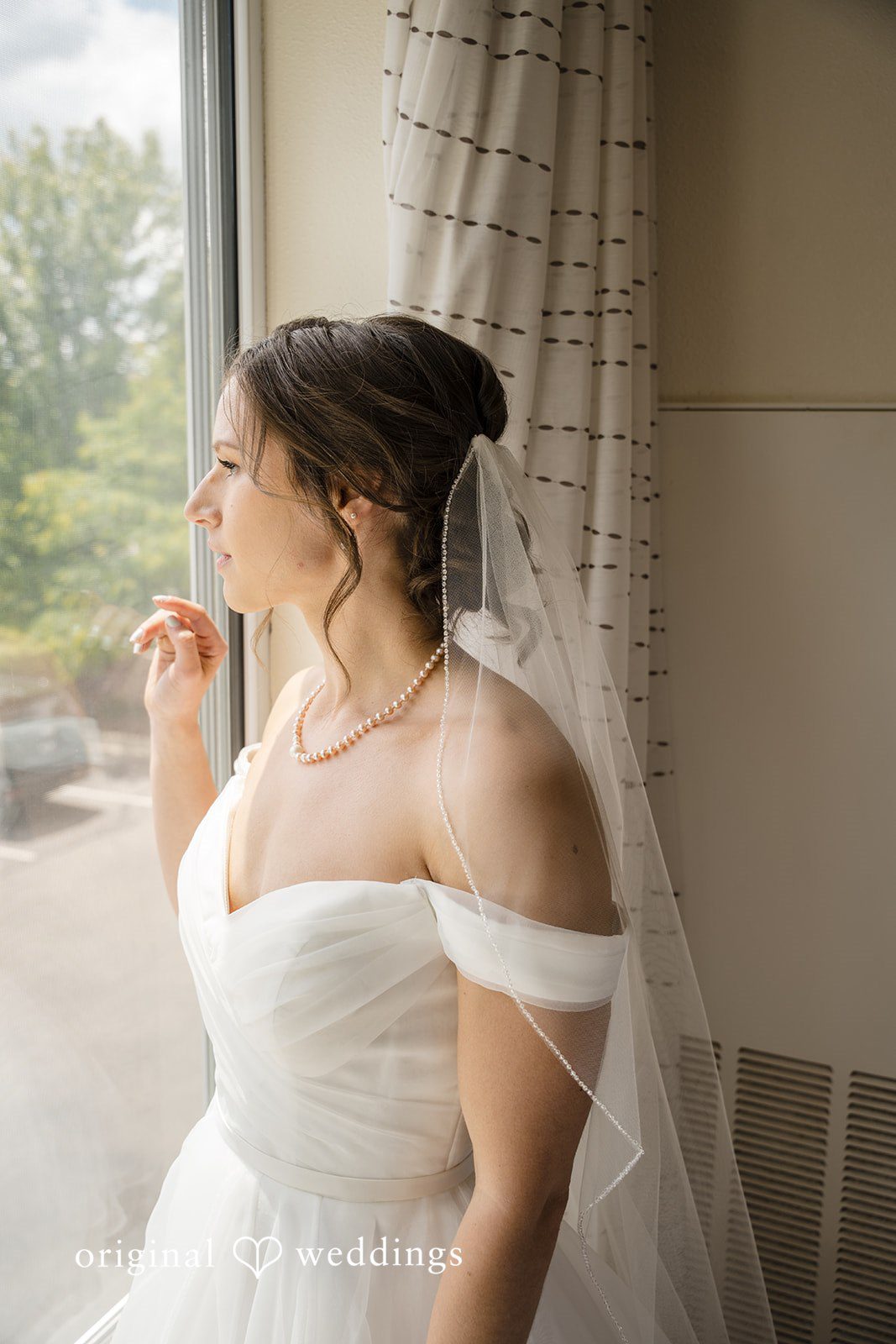 Emily + Maxwell A bride in an off-the-shoulder wedding gown looking out a window.