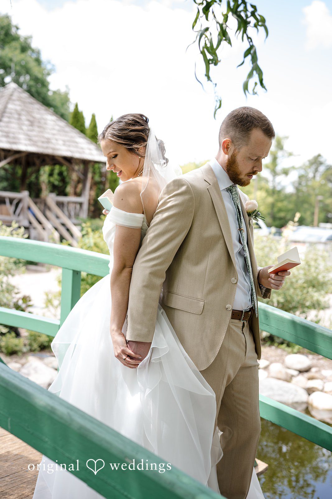 Emily + Maxwell A bride and groom standing back-to-back on a scenic wooden bridge walkway.