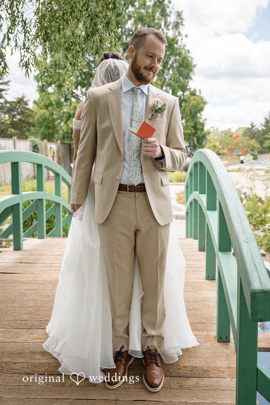 Emily + Maxwell A groom standing on an arched green bridge while his bride's gown flows around him.