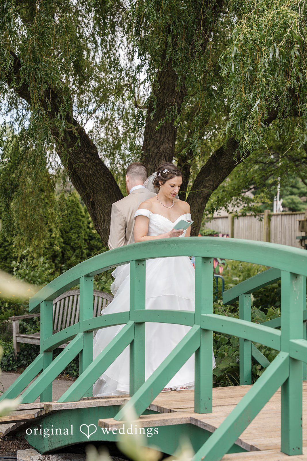 Emily + Maxwell A bride and groom standing together on an arched green bridge surrounded by trees.