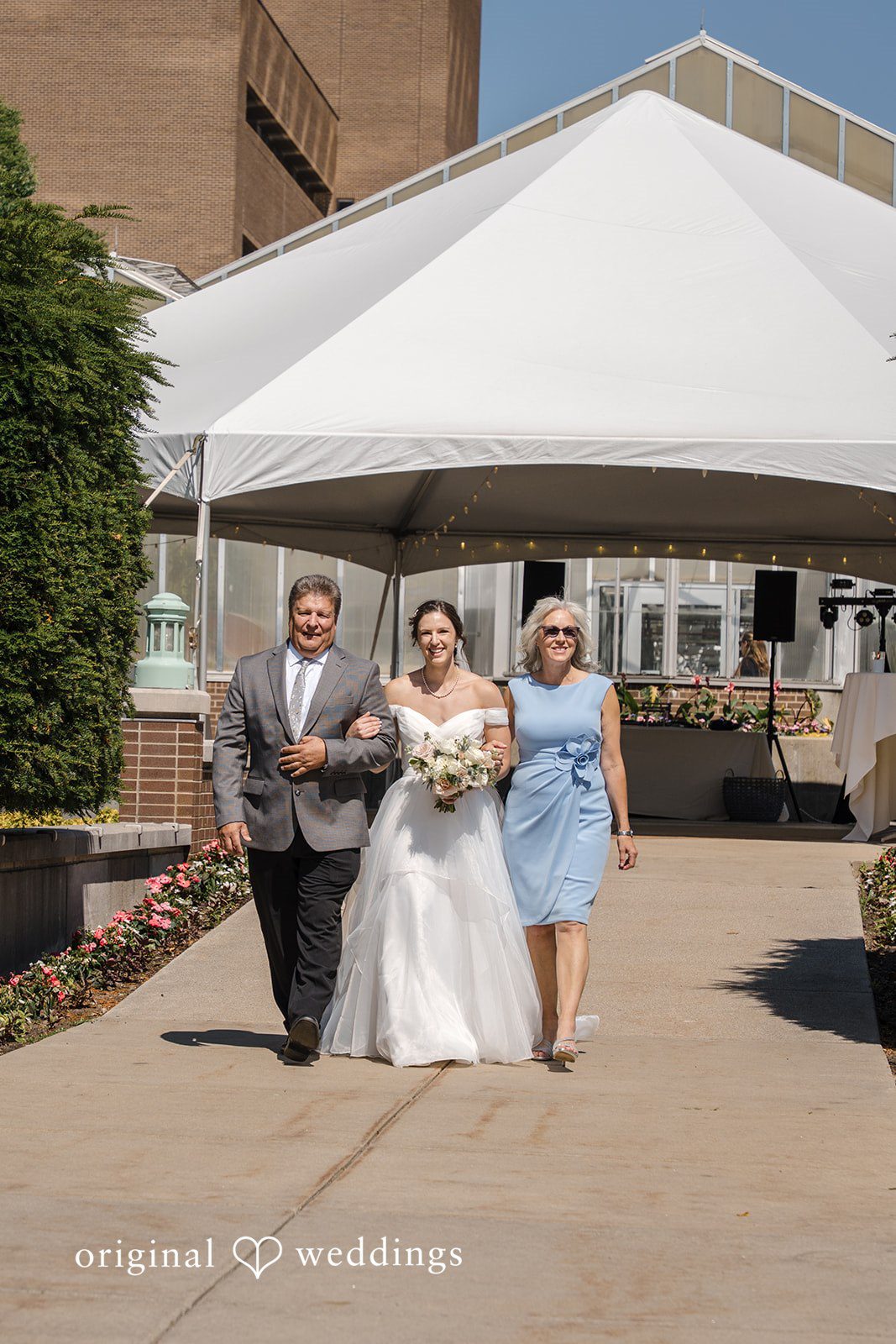 Emily + Maxwell A bride , man and women walking down a path under a white event tent.