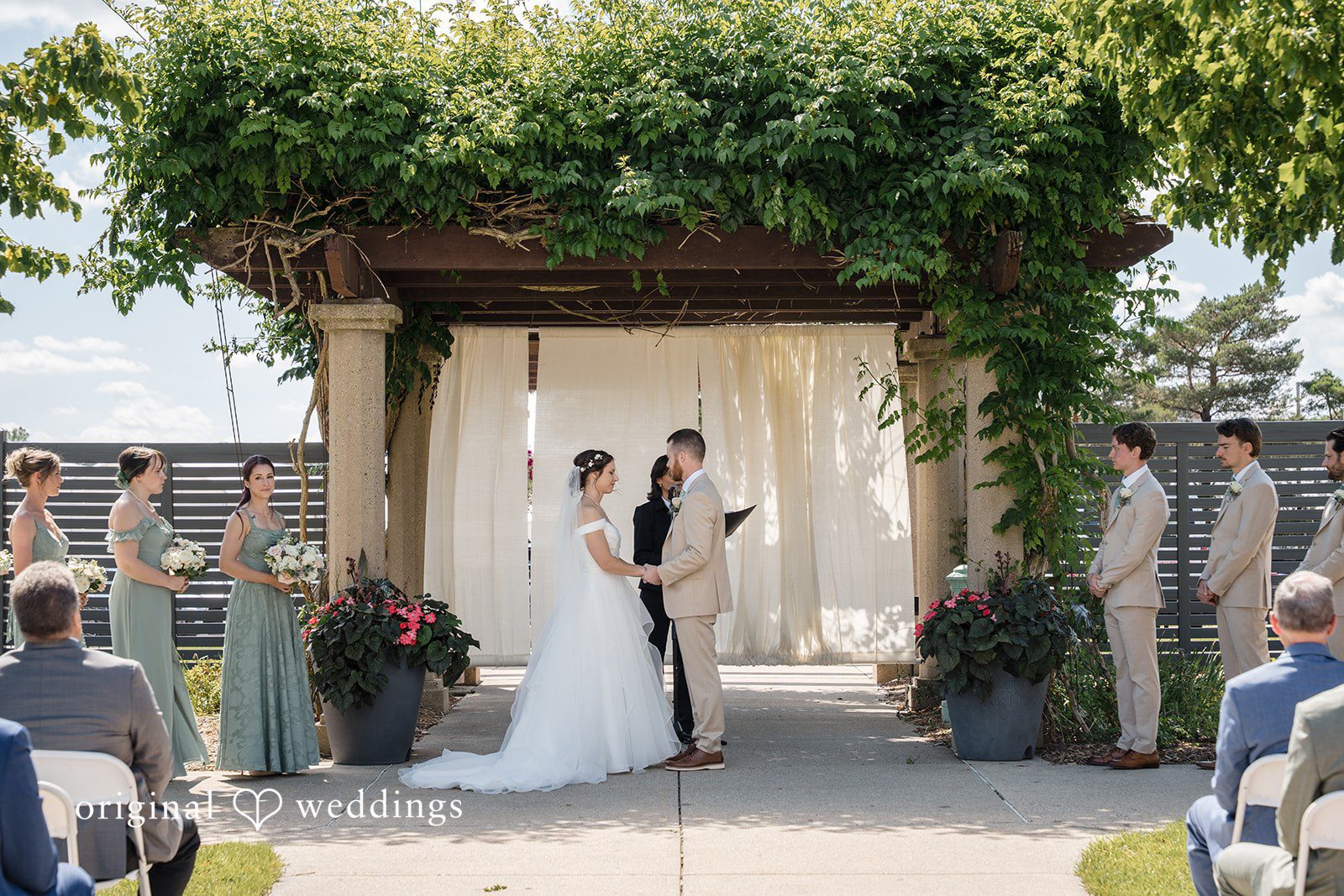 Emily + Maxwell A wide shot of a wedding ceremony under a lush green wooden pergola structure.