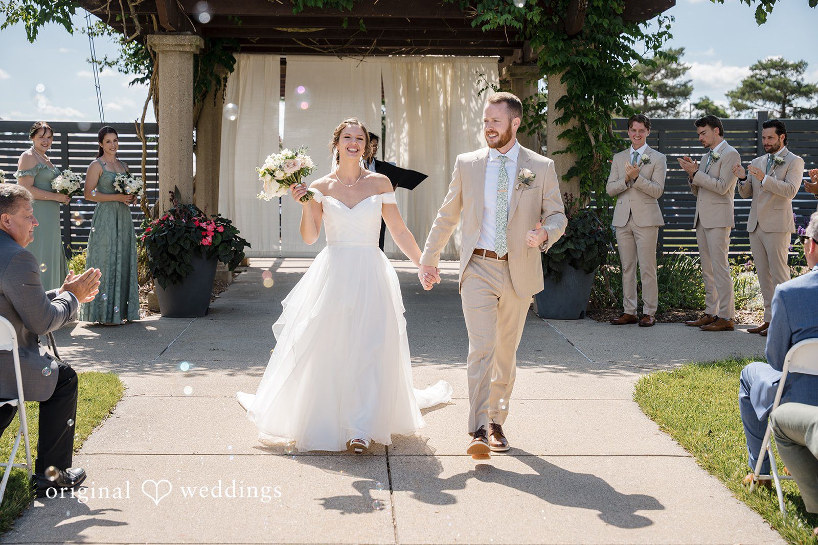 Emily + Maxwell A bride and groom holding hands and walking down a paved path as guests watch.