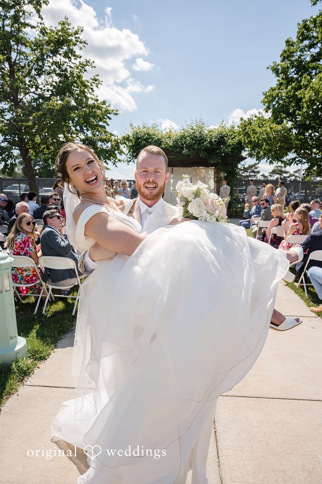 Emily + Maxwell A groom joyfully carrying his bride in his arms down a stone walkway.