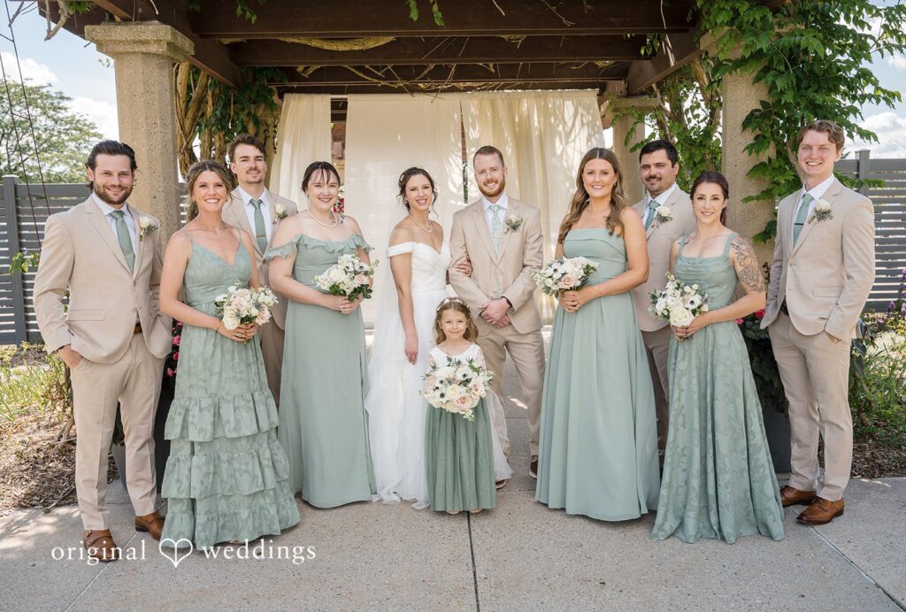 Emily + Maxwell A wedding party in sage green dresses and light suits posing together in a line at MSU Horticulture Gardens captured by Detroit Wedding Photographers from Original Weddings
