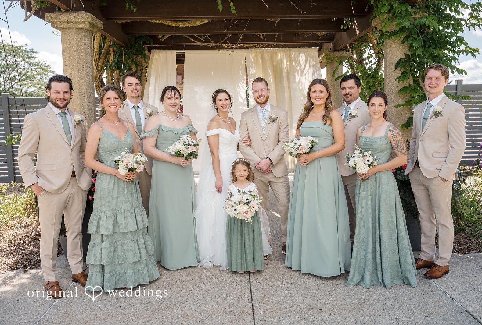 A wedding party in sage green dresses and light suits posing together in a line at MSU Horticulture Gardens captured by Detroit Wedding Photographers from Original Weddings