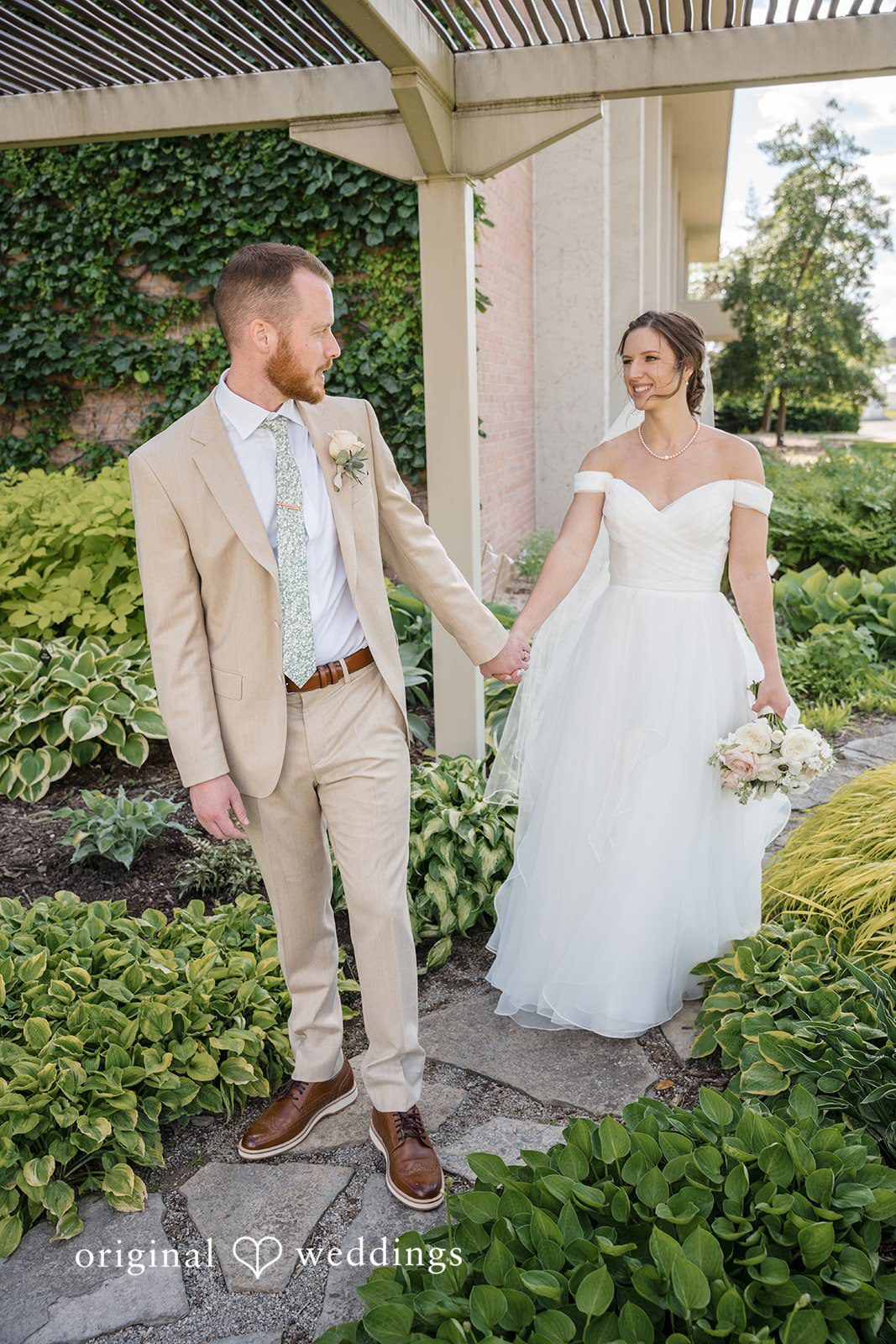 Emily + Maxwell A bride and groom holding hands and walking down a stone path next to a pergola.