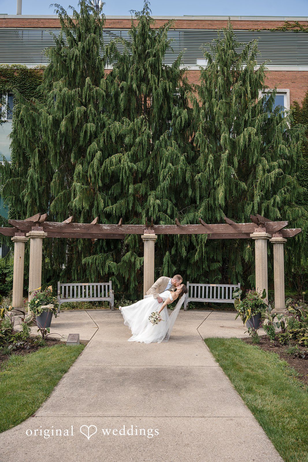 Emily + Maxwell A wide shot of a bride and groom posing together near a stone pergola structure.